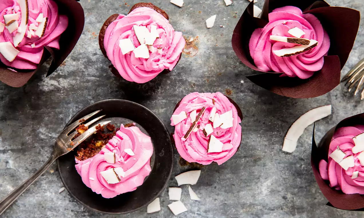 Randen-Mascarpone-Cupcakes mit leuchtend rosafarbener Cremehaube, garniert mit weißen Schokosplittern, in braunen Papierförmchen auf grauem Untergrund