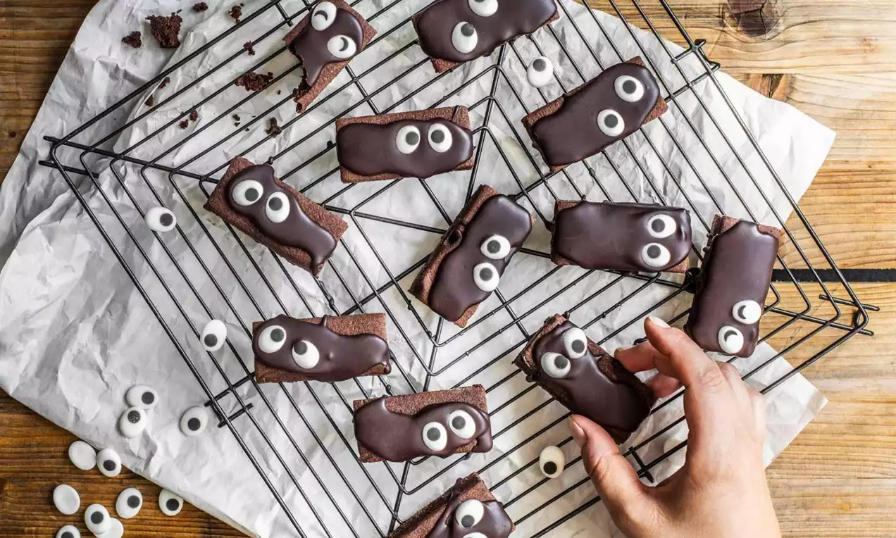 Rektangulære sjokoladebrownies med blank glasur og hvite sukkerøyne plassert på metallrist over bakepapir på trebord