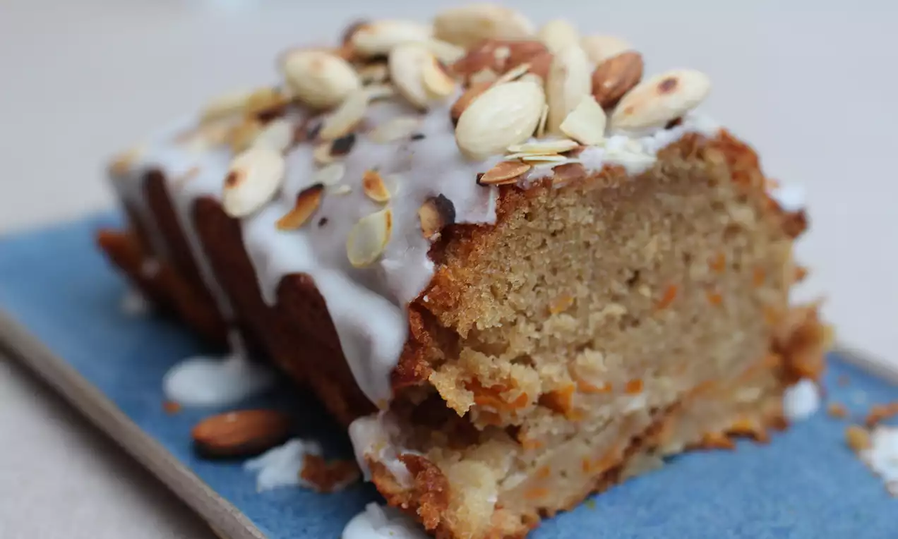 Butternutcake met lichtbruine kruimige structuur, witte glazuurlaag en amandelen, geserveerd op een blauw bord