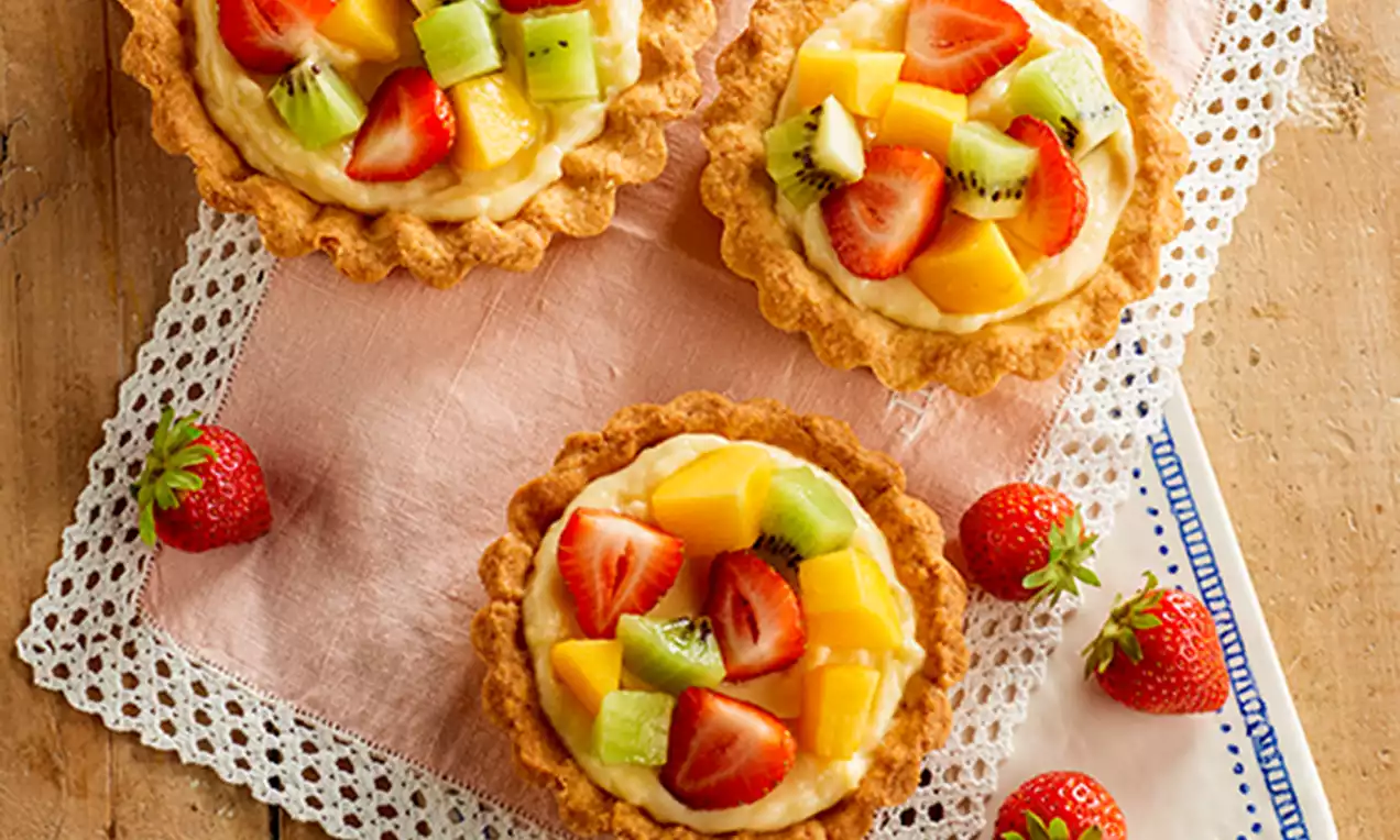 Tartaletas de masa dorada con crema suave, cubiertas de trozos brillantes de kiwi, mango y fresas sobre un paño rosa y encaje blanco