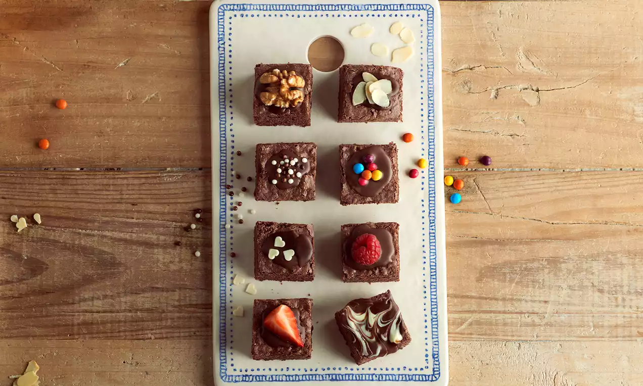 Cuadrados de brownie oscuro con coberturas variadas como nueces, frambuesa, fresas, almendras y pequeños dulces de colores en una bandeja blanca rectangular con borde azul sobre una mesa de madera clara