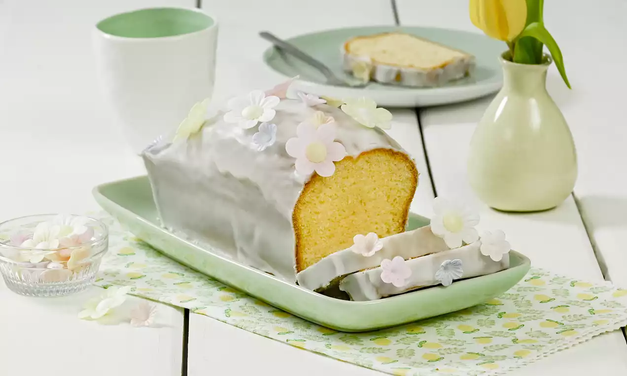 Plumcake giallo soffice ricoperto di glassa bianca lucida e decorato con fiori di zucchero color pastello su un piatto verde rettangolare