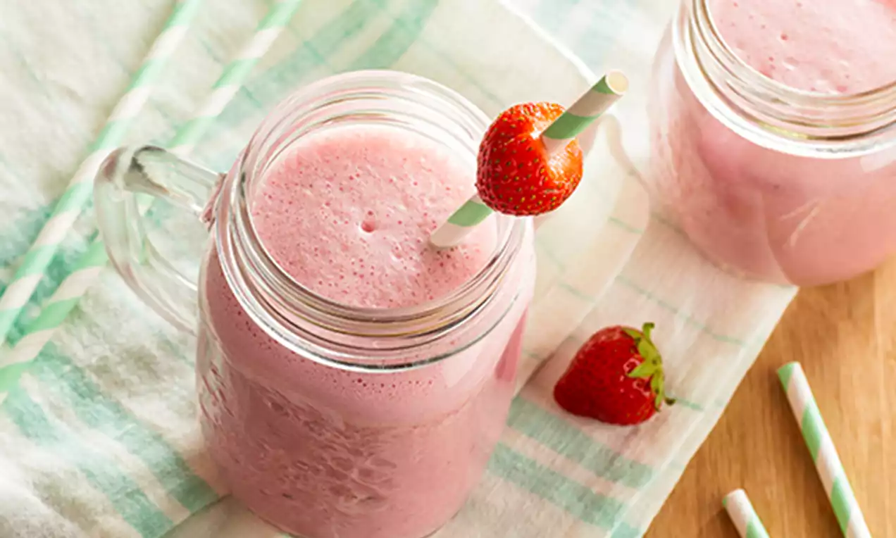 Batido rosa cremoso en jarra de cristal con pajita rayada verde y blanca, decorado con una fresa y dispuesto sobre un paño claro doblado