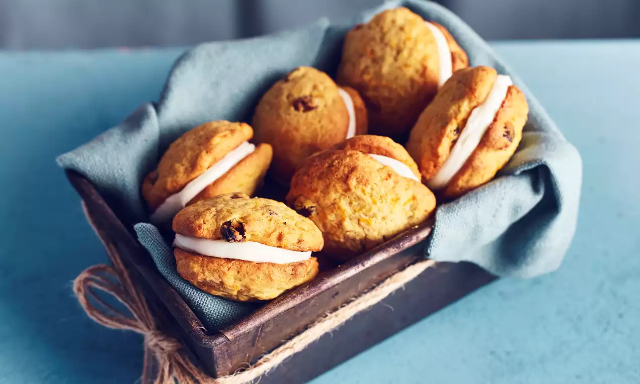 Golden-brown carrot cakes with soft, rounded edges sandwiching white cream filling, arranged in a dark tray lined with blue fabric
