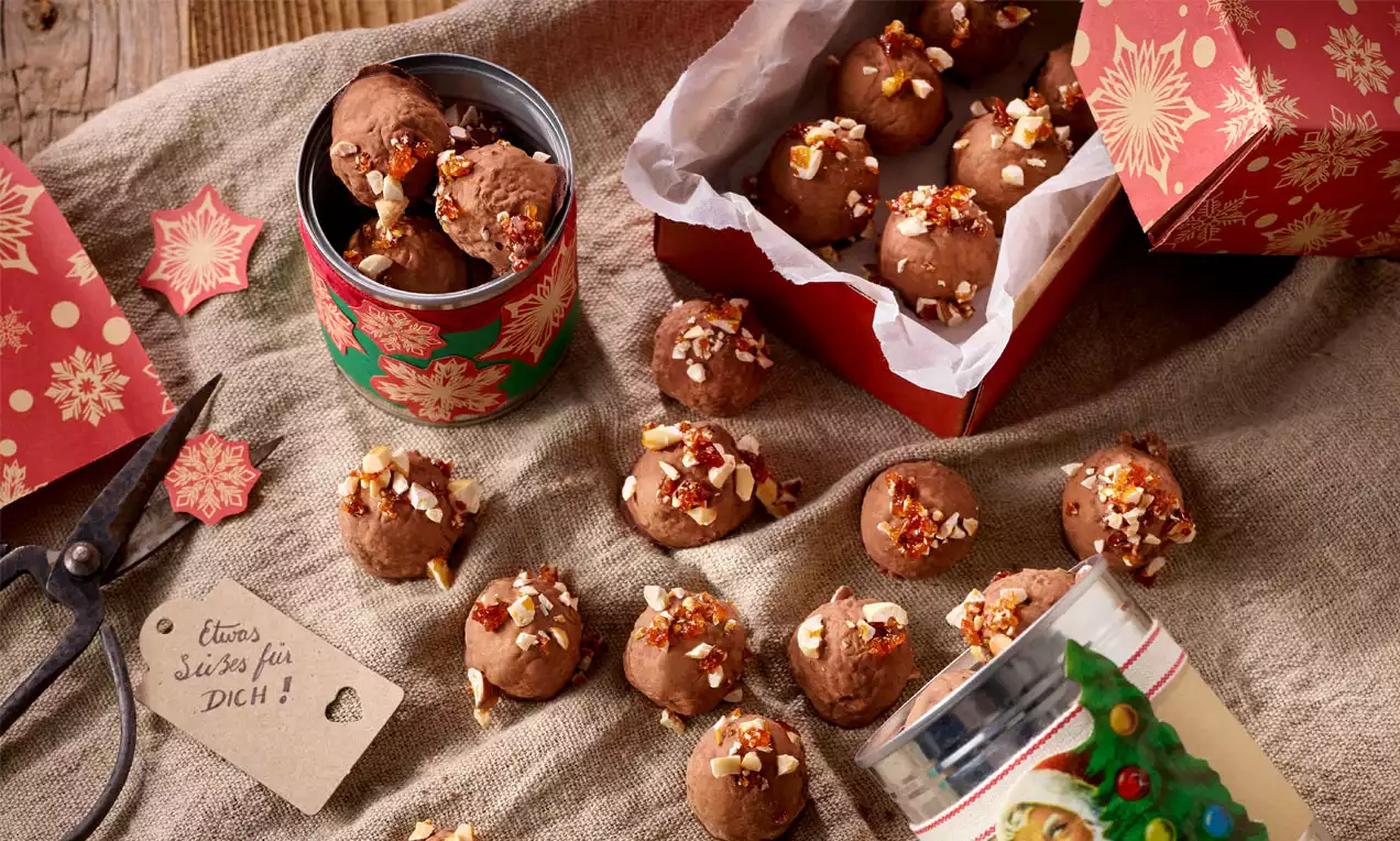 Runde Pralinen aus dunkler Schokolade, bestreut mit krokantem Nussstückchen, liegen teils in festlichen roten Geschenkboxen auf beigem Stoff