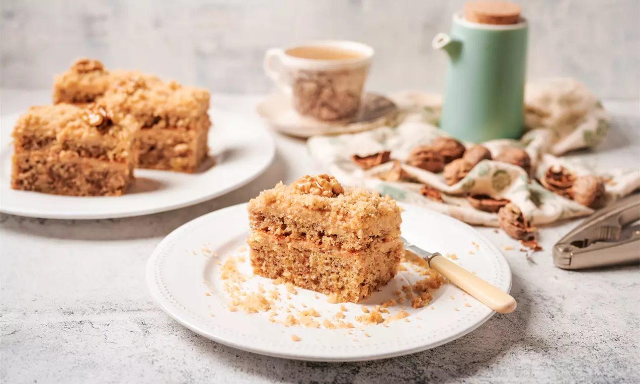 Квадратна орехова паста със златисто-кафяв цвят и ронлива текстура върху бяла чиния, заобиколена от орехи и ножче с дървена дръжка