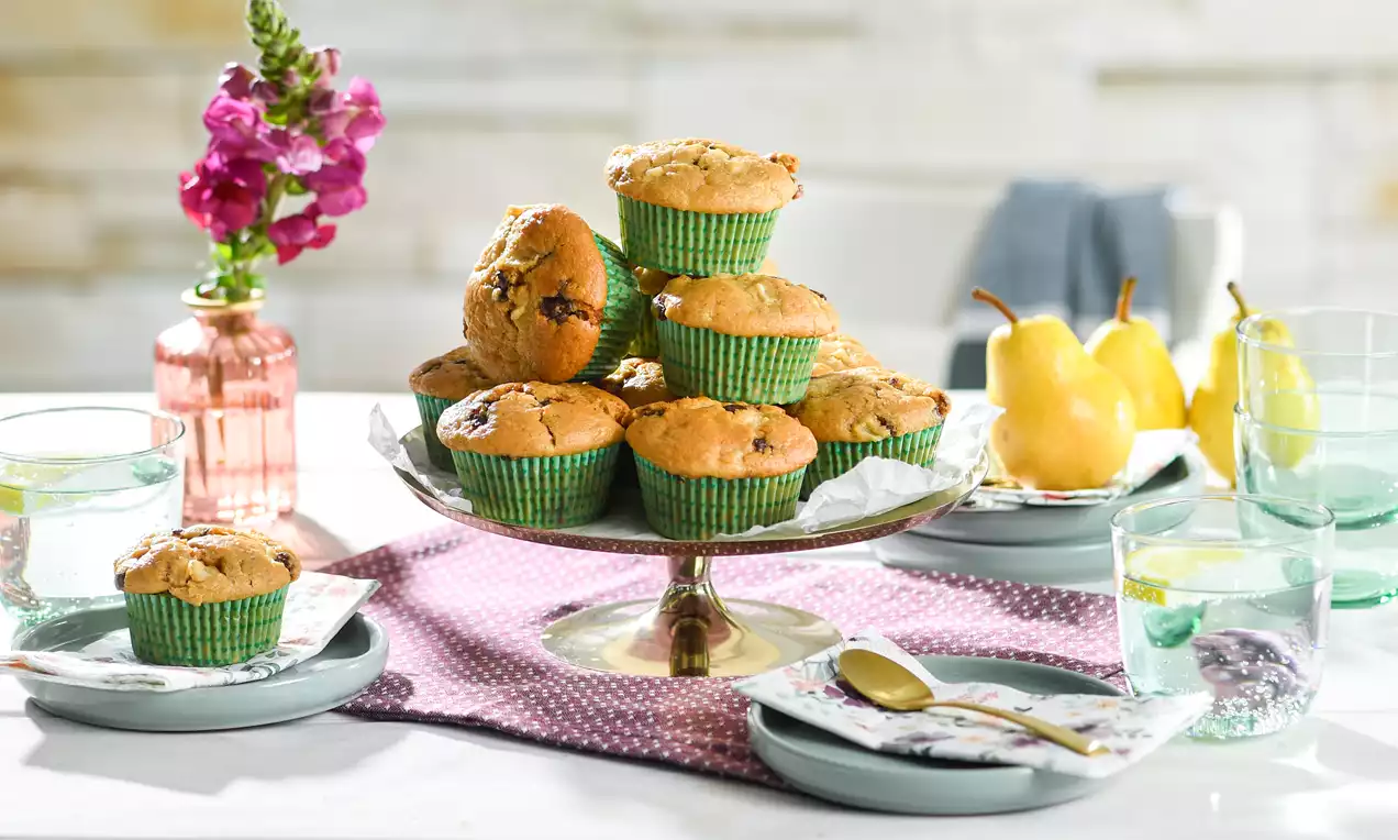 Goldbraune Birnenmuffins in grünen Papierförmchen auf einer Glas-Etagere, umgeben von gelben Birnen und gedecktem Tisch mit pastellfarbenem Geschirr