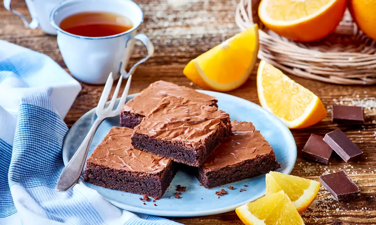 Kwadratowe kawałki czekoladowego brownie o gładkiej, błyszczącej powierzchni na jasnym talerzu, obok plasterków pomarańczy i kawałków czekolady