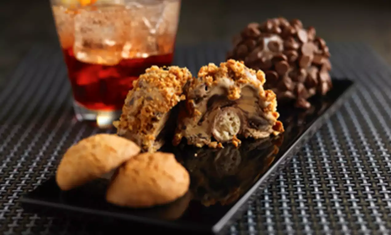 Tartufo with a textured chocolate shell, sliced to reveal layers of cream and filling, served on a black plate with biscuits and a drink