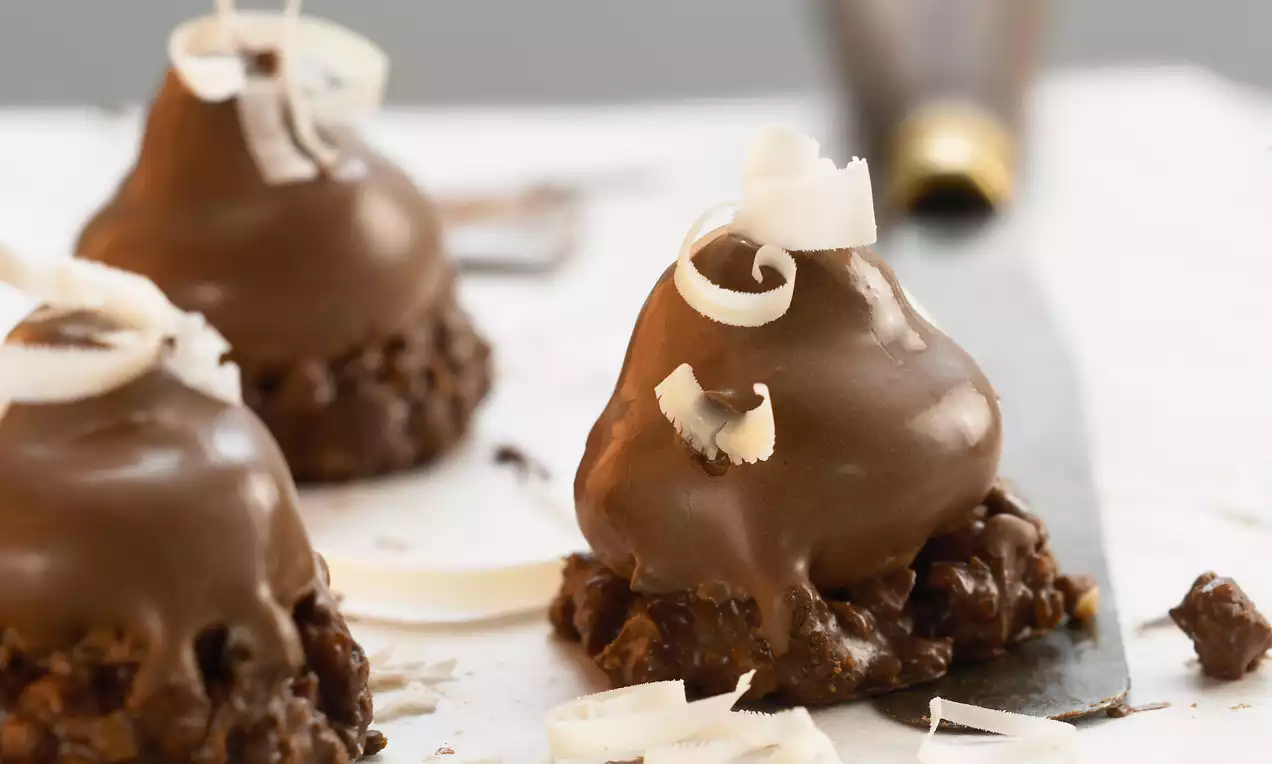 Petits monticules enrobés de chocolat couleur moka sur une base croquante, garnis de fins copeaux de chocolat blanc et disposés sur une surface claire