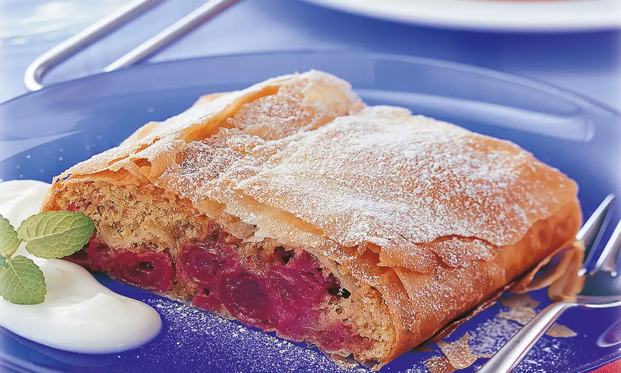 Rechteckiger Weichselstrudel mit goldbrauner, knuspriger Teighülle und staubzuckerweißem Topping, daneben weiße Schokosoße auf blauem Teller