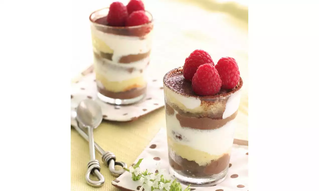 Layered mini trifles in clear glasses with chocolate, vanilla cream, and sponge, topped with red raspberries on dotted napkins