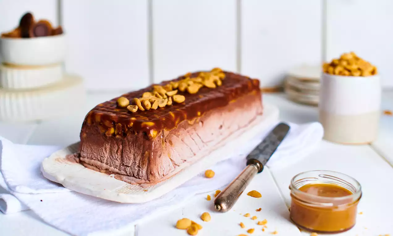 Rechteckiges Schokoeis mit glatter Oberfläche, überzogen von glänzender Karamellschicht und gerösteten Erdnüssen auf weißer Platte