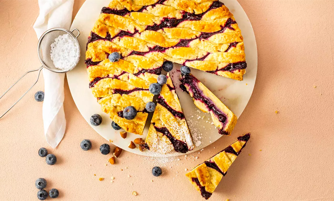 Боровинков пай с кръгла форма и златисто тесто с кръстосани ленти, гарниран със свежи боровинки върху светла плоска чиния