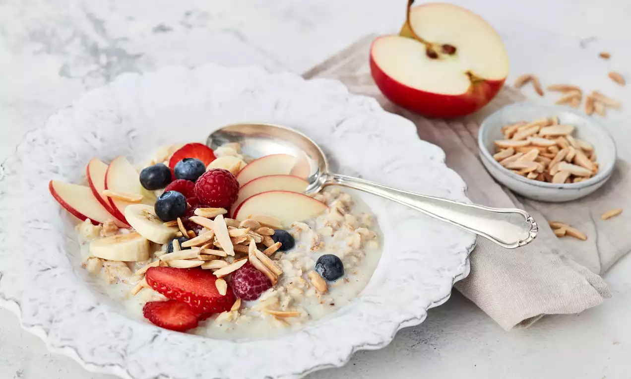 Porridge mit cremigen Haferflocken, roten Beeren, Bananenscheiben, einem Löffel Joghurt und Honig, angerichtet in weißer Schale auf Holz