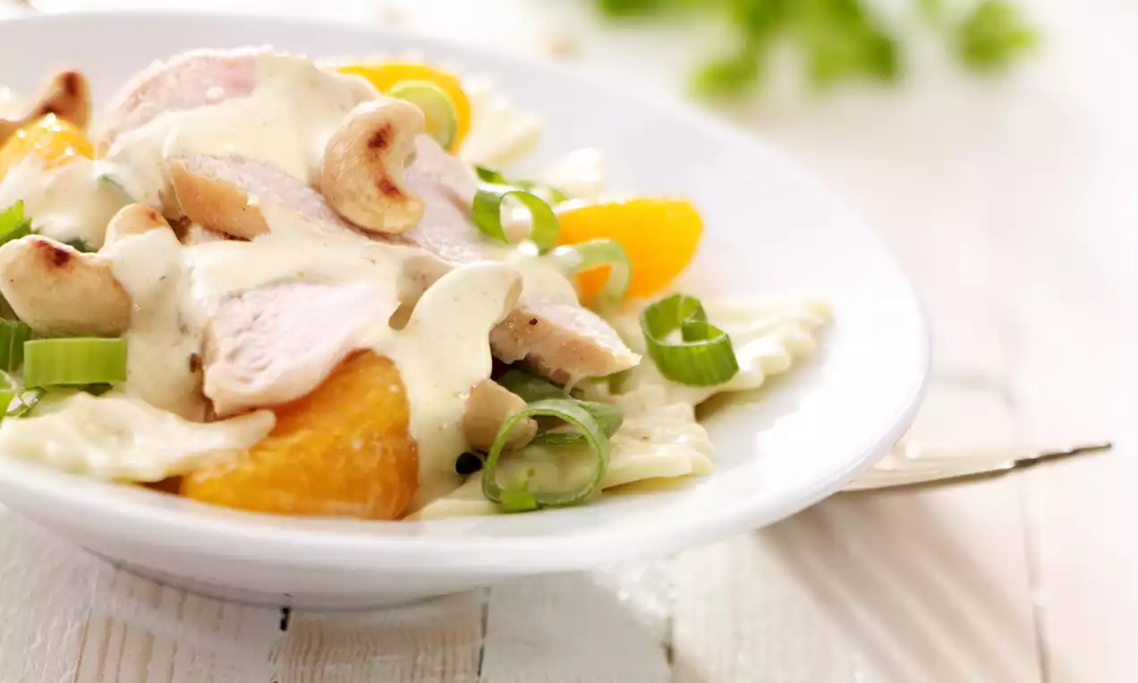 Nudelsalat mit hellen Teigwaren, cremiger Sauce, Hähnchenstücken, orangefarbenen Mandarinen und grünen Frühlingszwiebeln auf weißem Teller