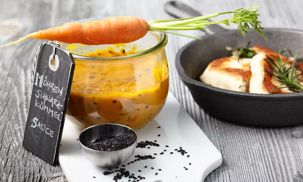 Goldorange Möhrensoße mit schwarzem Kümmel in einem Glas, dekoriert mit einer frischen Karotte, neben einer Pfanne mit Käse auf Holzbrett
