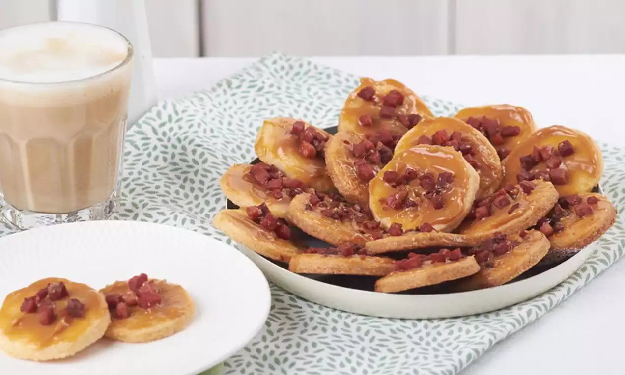 Cookies dorés et croustillants ovales nappés de caramel ambré et éclats de bacon rouge sombre sur une assiette blanche posée sur un tissu imprimé