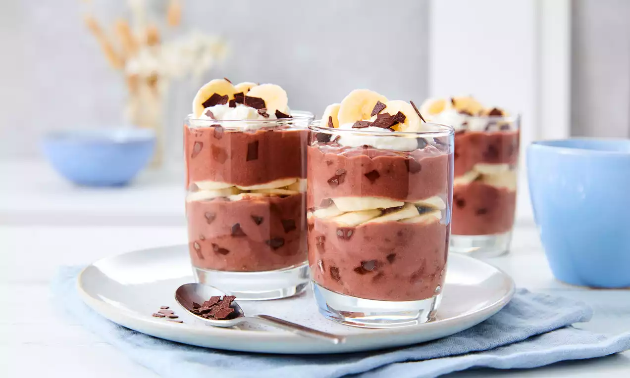 Schoko-Bananen-Dessert in Gläsern, mit dunkelbrauner Creme, frischen Bananenscheiben, Sahnehaube und Schokoraspeln auf weißem Teller