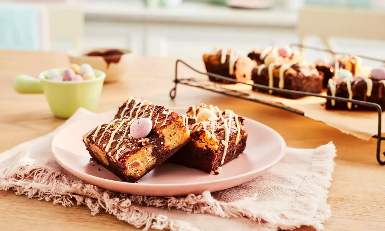 Brownies moelleux couleur cacao ornés de mini-œufs pastel et de filaments de chocolat blanc sur une assiette rose posée sur un tissu