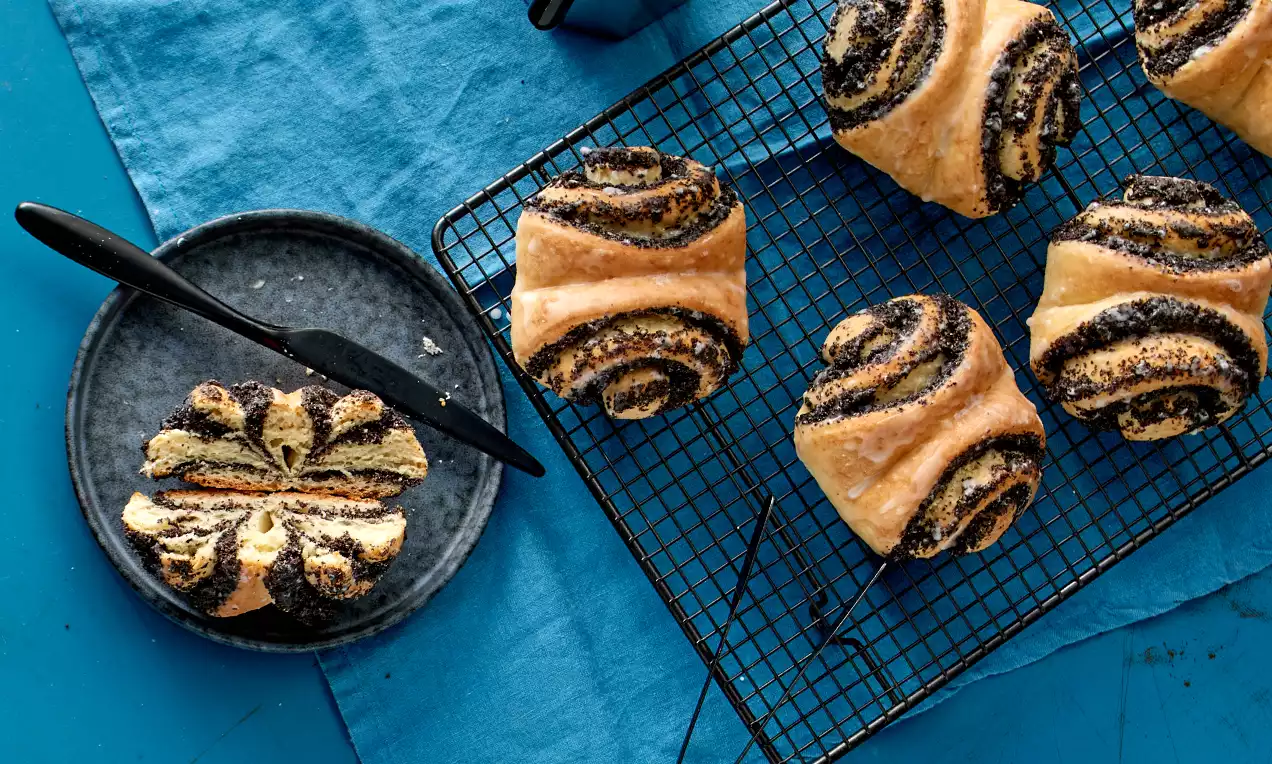 Goldbraune Mohn-Franzbrötchen mit spiralförmigen Streifen aus dunkel glänzendem Mohn auf einem schwarzen Gitter und blauem Tuch arrangiert