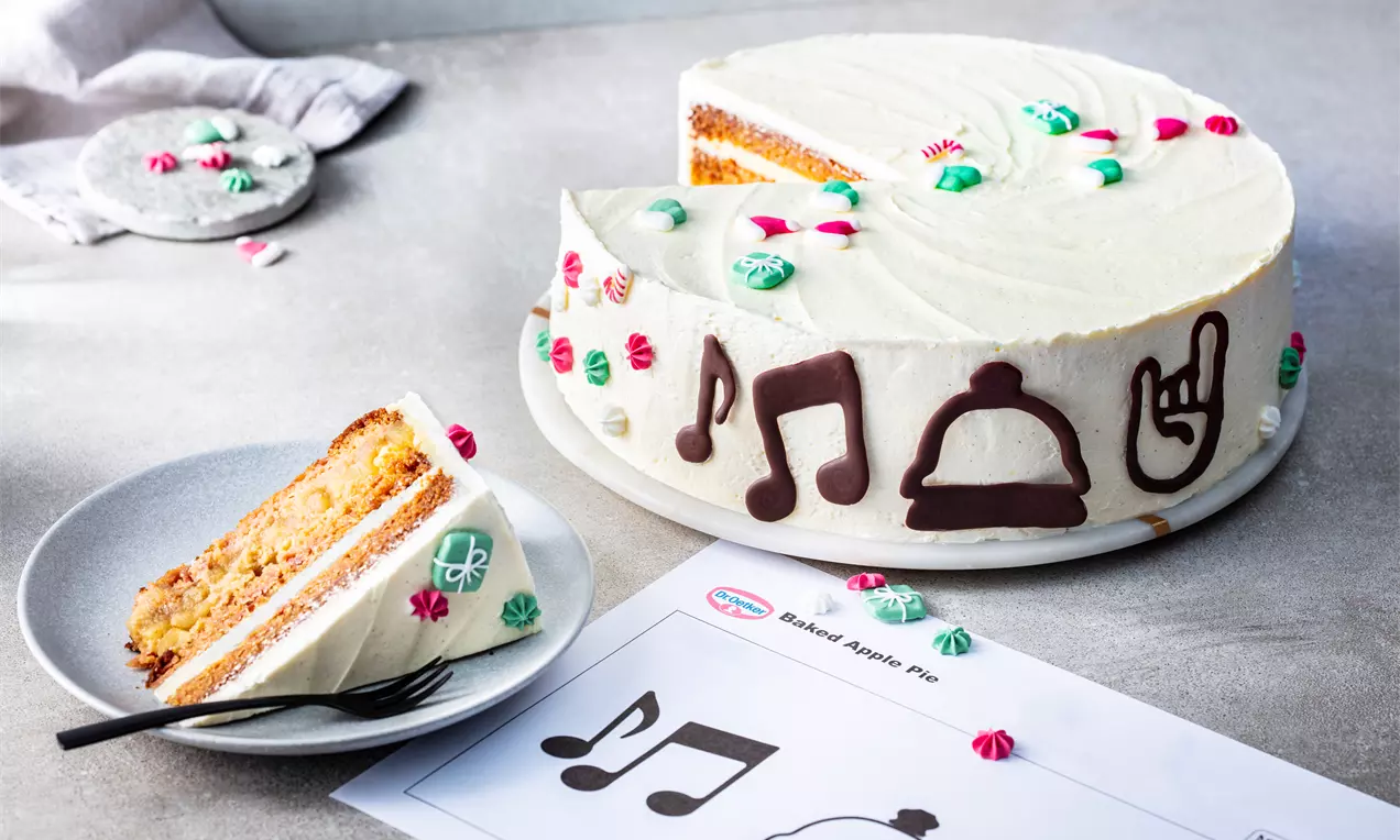 Кръгла ябълкова торта с бял крем, свързан с флорални декорации и шоколадови фигури, върху сиво плато с рязано парче до нея