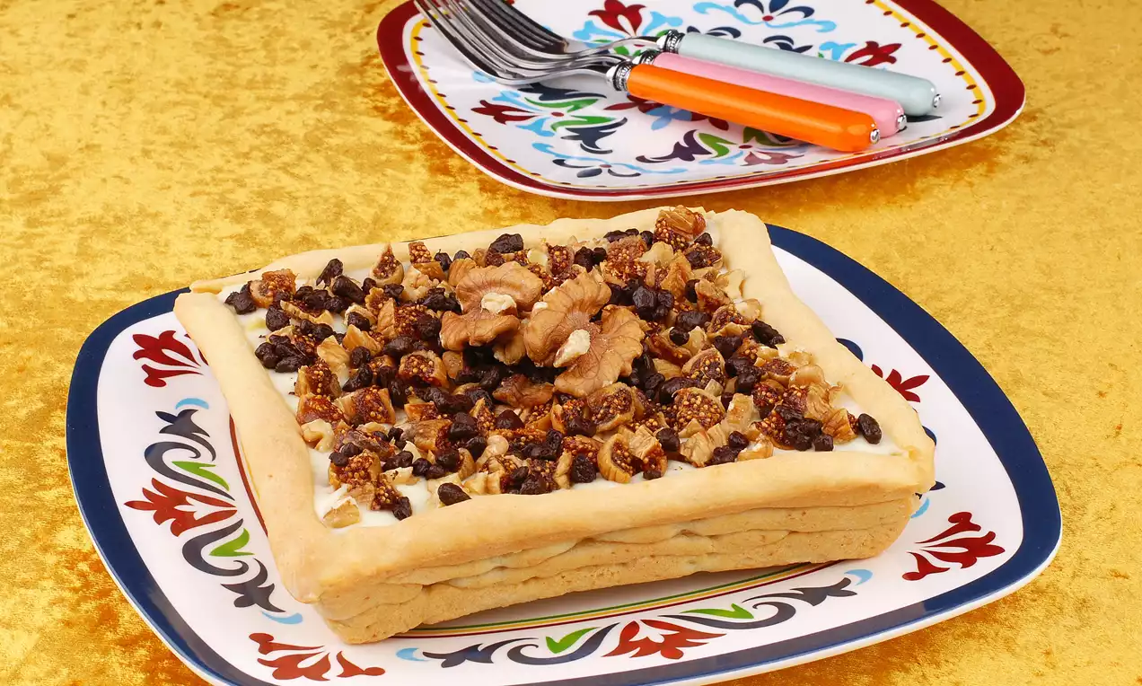 Dikdörtgen bir tabakta ince hamur tabanlı tart, beyaz kremayla kaplı ve üzerine ceviz, kuru incir ile çikolata parçaları serpilmiş