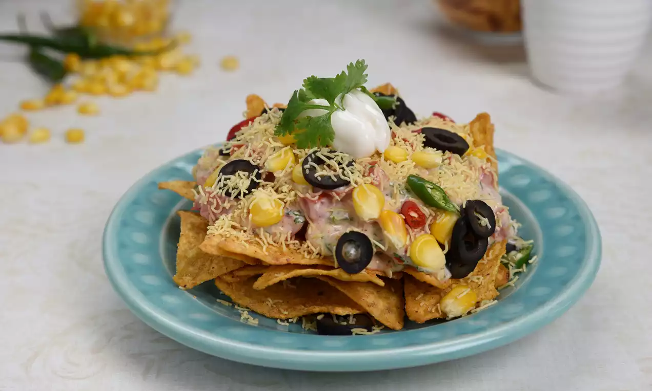 Nacho chips layered with corn, diced vegetables, black olives, grated cheese, creamy white dollop and green coriander on a turquoise plate