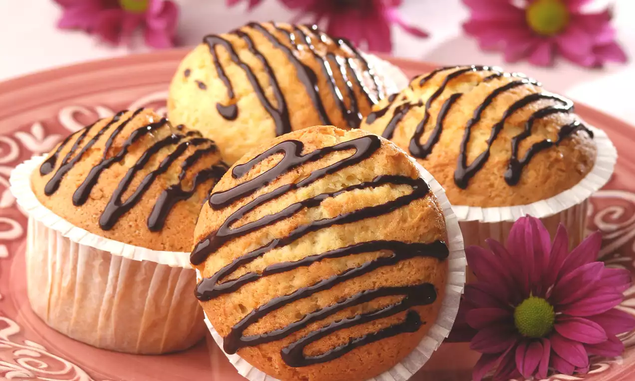Pembe desenli tabakta çikolata sosuyla çizgilerle süslenmiş altın sarısı muffinler, kenarında pembe kasımpatı çiçekleriyle