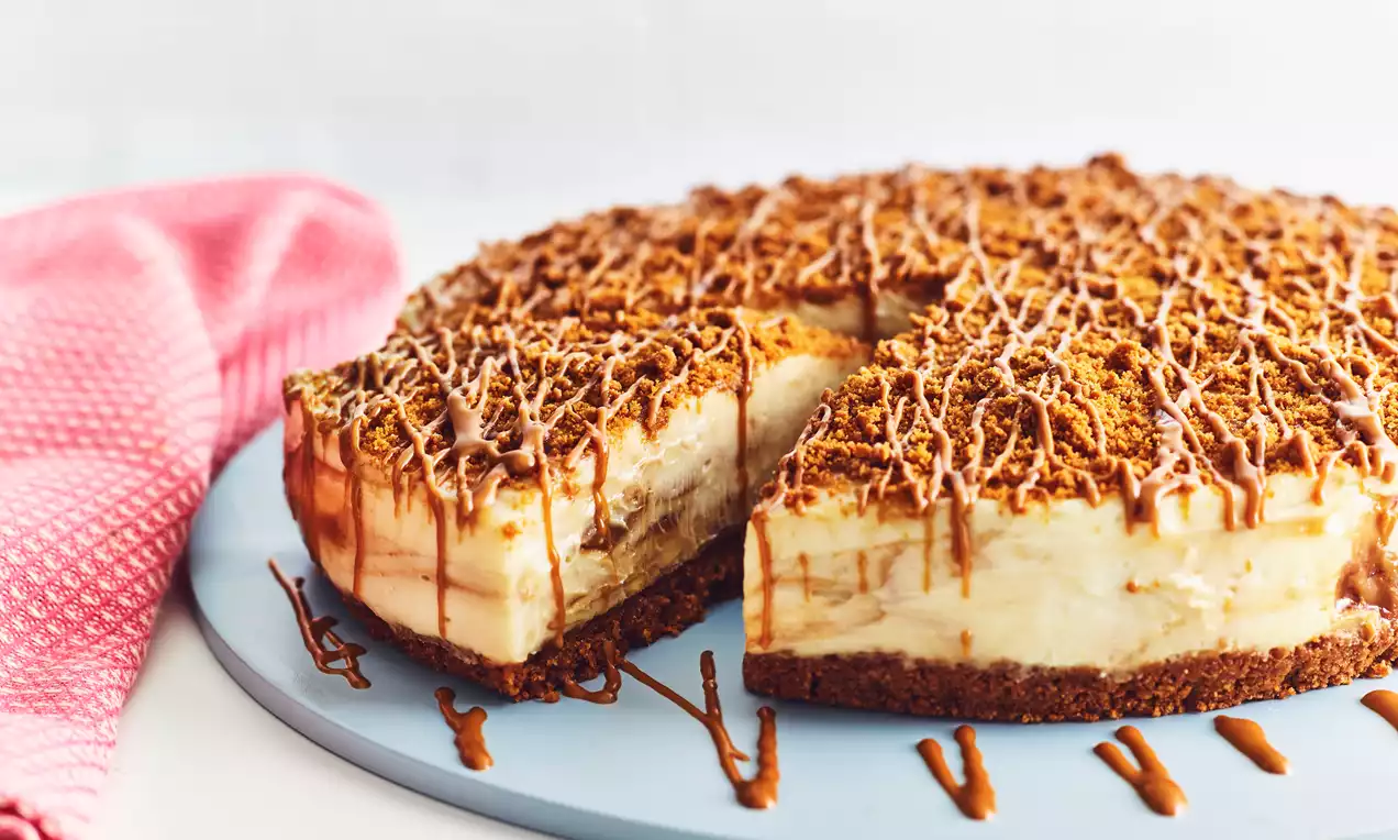 Round cheesecake with a crumbly brown biscuit base and creamy off-white filling topped with crushed Biscoff and caramel drizzle on a pale blue plate