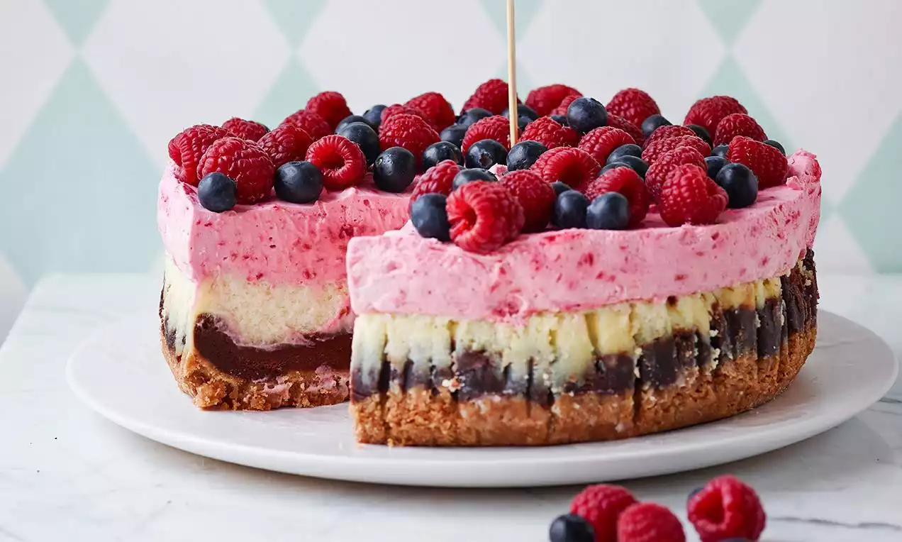 Rund ostekake med gylden kjeksbunn, lag av sjokolade og vaniljefyll toppet med rosa bærmousse, friske bringebær og blåbær på hvitt fat