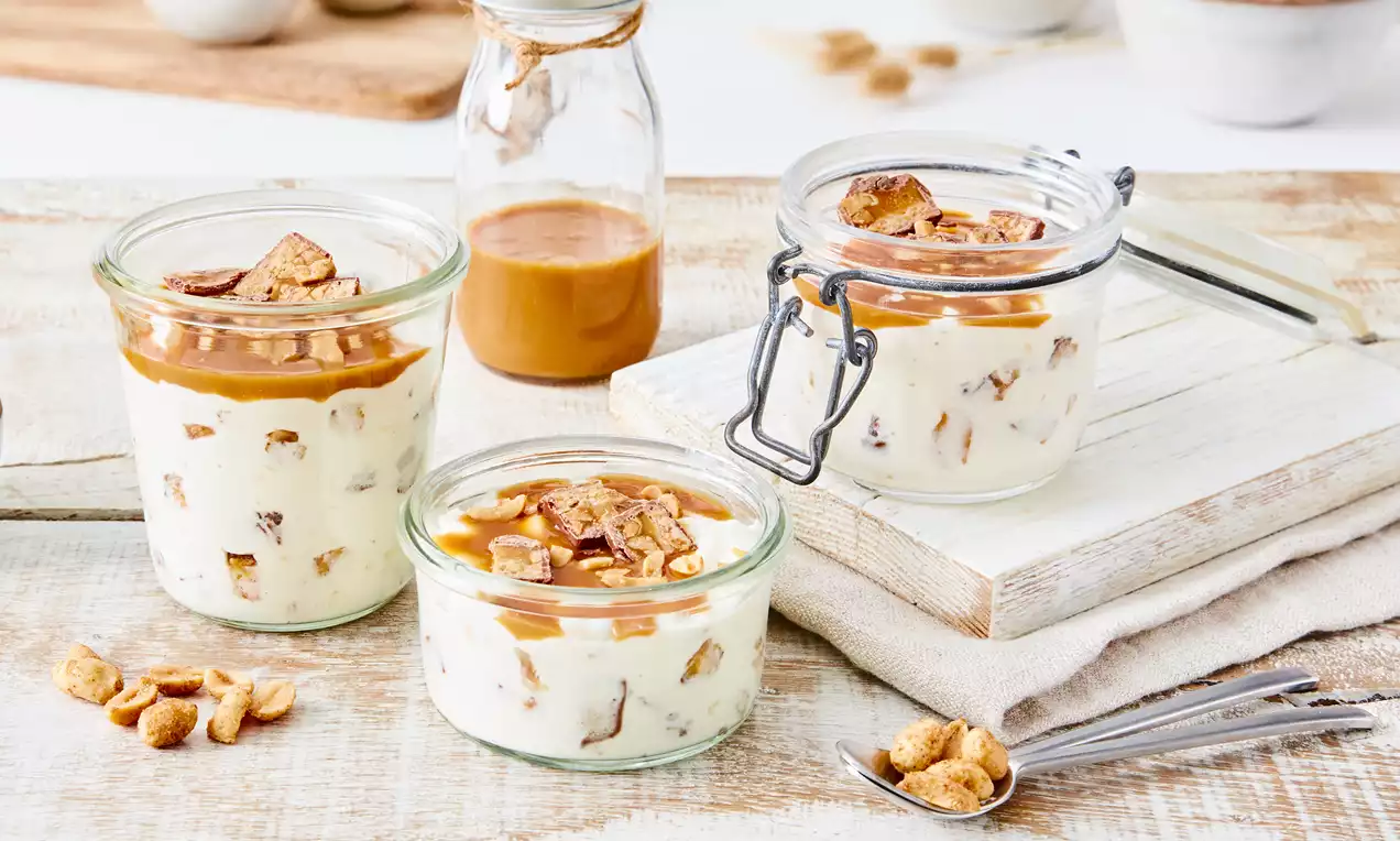 Cremiges Dessert in Glasbehältern mit goldbrauner Karamellsoße, stückiger Schokoladenfüllung und Erdnüssen auf hellem Holzbrett angerichtet
