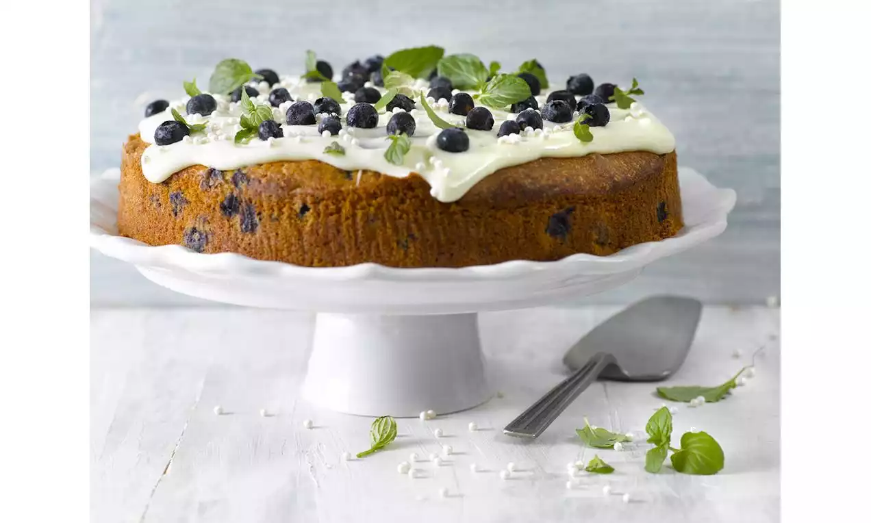 Rund blåbærkake med gyllenbrun skorpe, hvit glasur, friske blåbær og mynteblader på kakefat med kakespade og grønne blader rundt