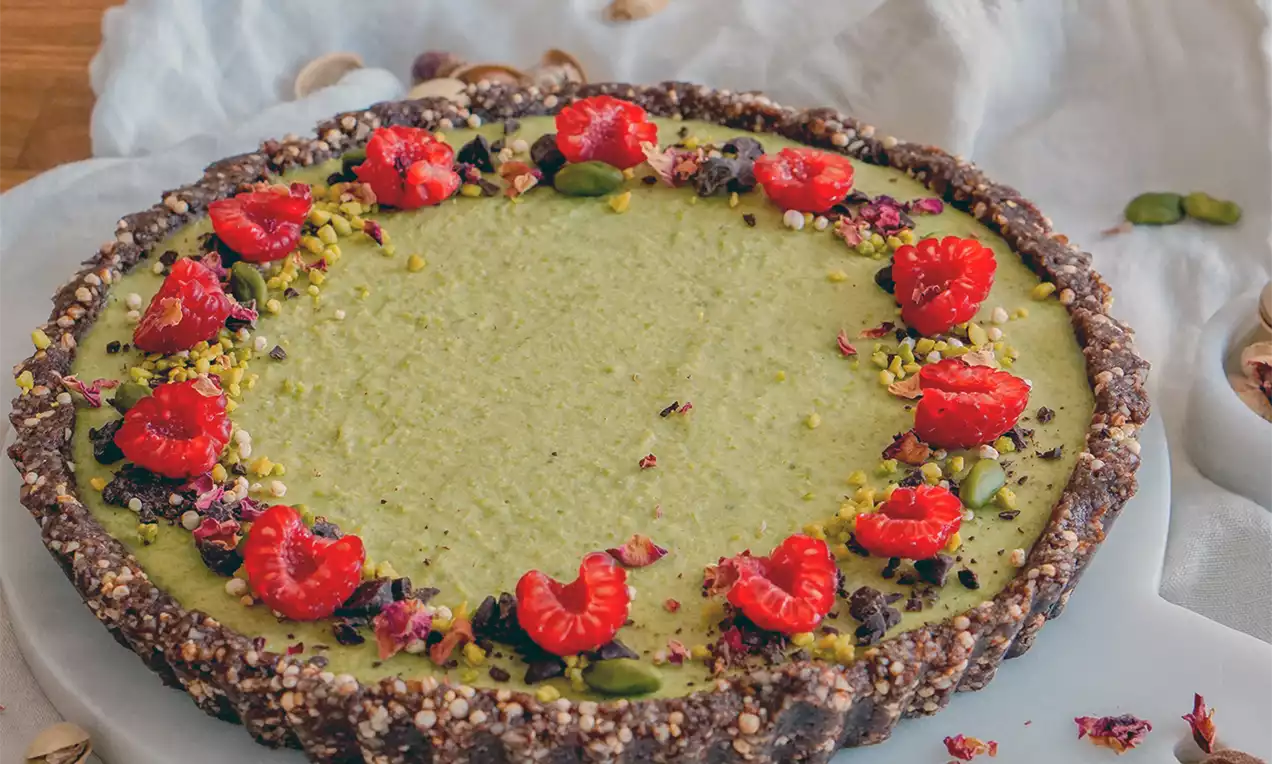 Runde Pistazien-Tarte mit grünem Belag, dunklem Nussboden, dekoriert mit frischen Himbeeren, Pistazienstücken und Blütenblättern auf weißer Platte