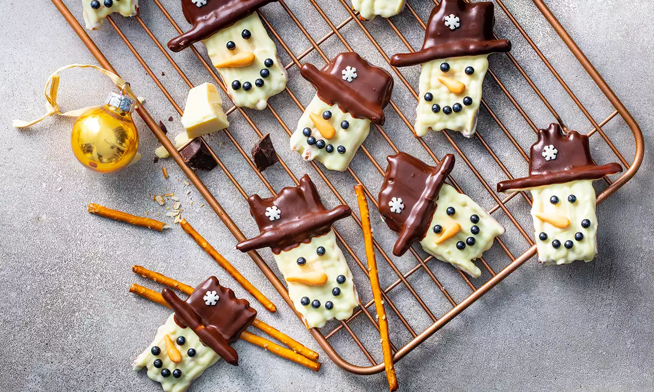 Snövit glasyr på kakor formade som snögubbar med chokladhattar, svarta dekorationsögon och näsor på ett galler med kringlor och julkula