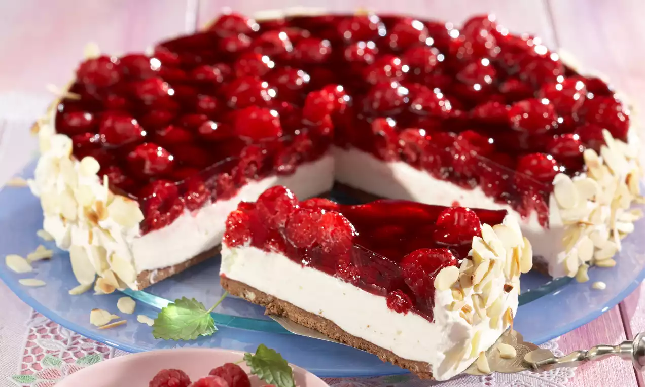 Sommerliche Torte mit hellem Sahneboden, glänzendem rotem Himbeerspiegel, gerösteten Mandelblättchen und frischen Himbeeren auf blauem Teller