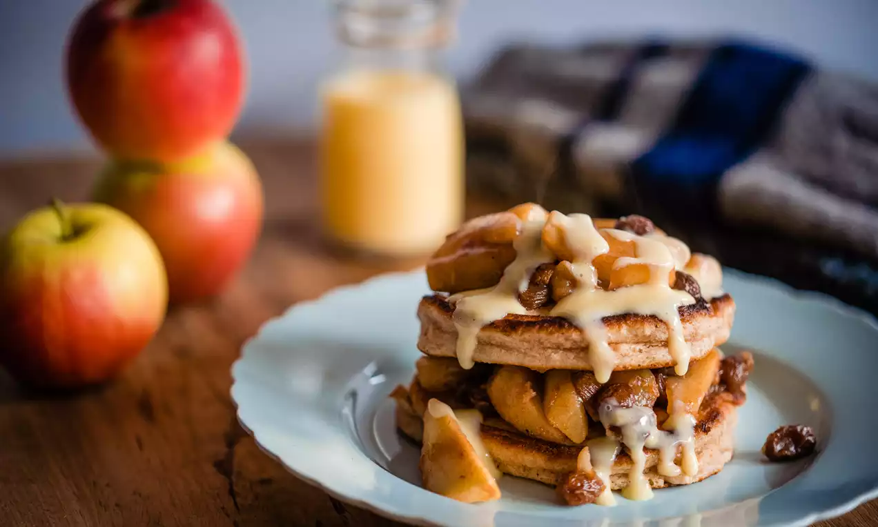 Drie stapels goudbruine pannenkoeken met gekaramelliseerde appelpartjes, rozijnen en romige vanillesaus op een wit bord, met appels en een glazen flesje saus op een houten tafel