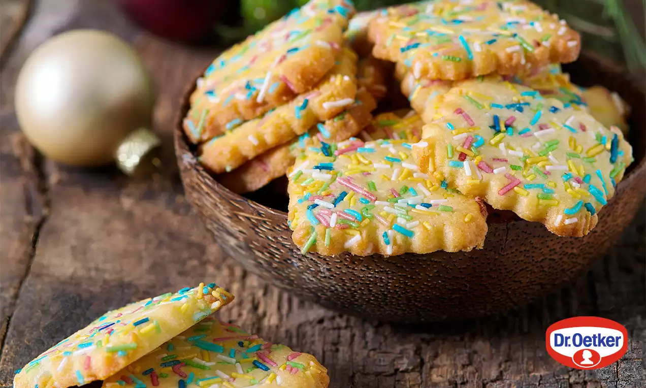 Máslové sušenky se zubatými okraji, světle zlatavé, posypané barevnou cukrovou polevou, uspořádané v hnědé misce na dřevěném povrchu