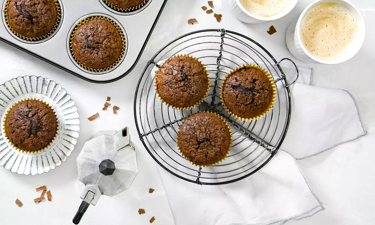 Saftige Brownie-Muffins mit goldenen Förmchen, dunkelbrauner Oberfläche und zarter Kruste auf einem Kuchengitter neben Cappuccinos und Mokkanne