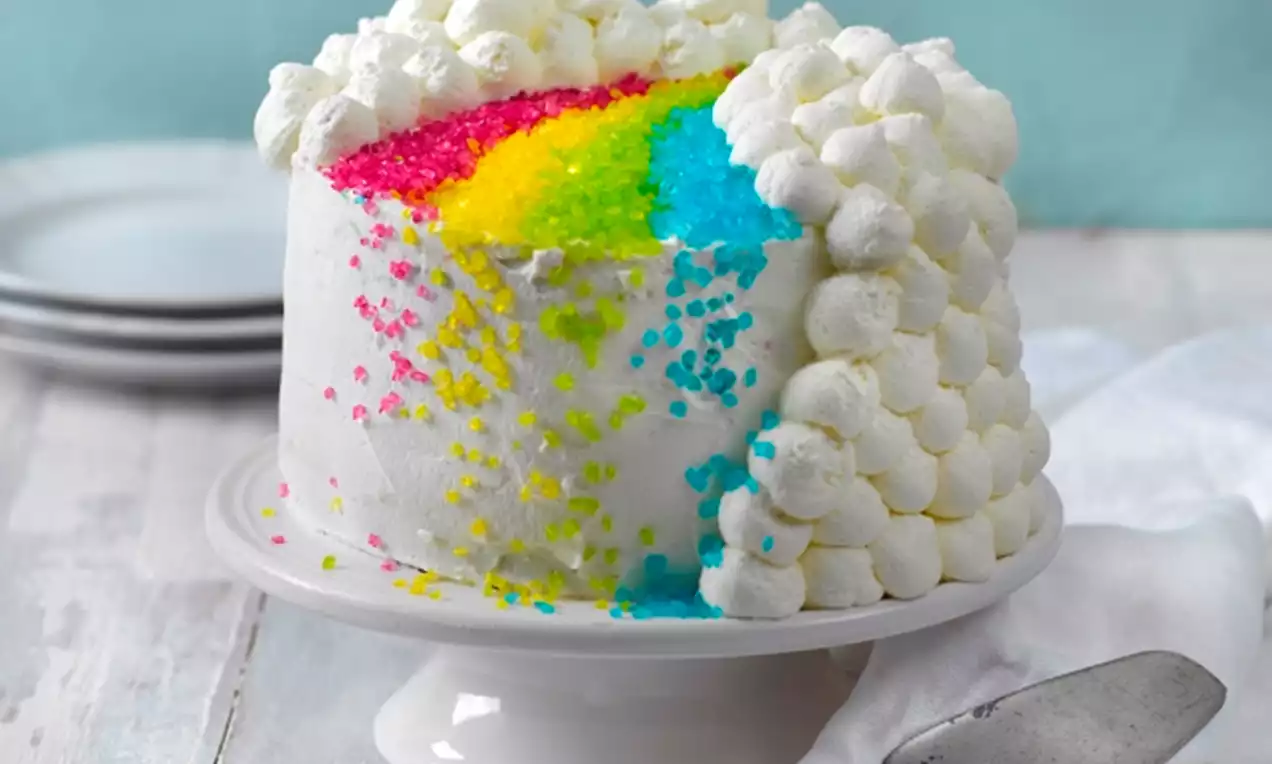 Sateenkaaren kirkkain värein täytetty korkea valkoinen kakku, pehmeästi kermavaahtopursotettu, värikkäillä sokerirakeilla koristeltu