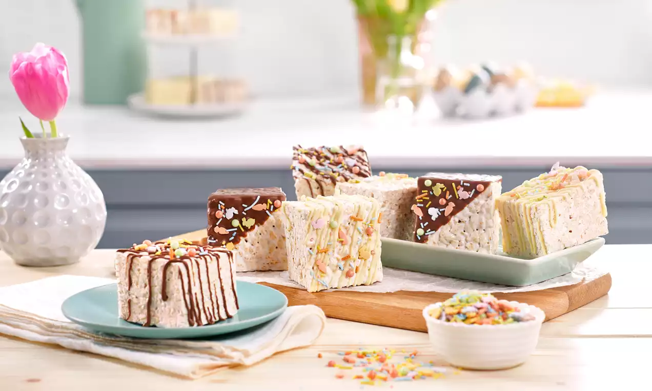 Bunte, dekorierte Reiscrackerschnitten in Würfelform auf Holzbrett und grünem Teller neben einer Schale mit bunten Puffreis-Stückchen