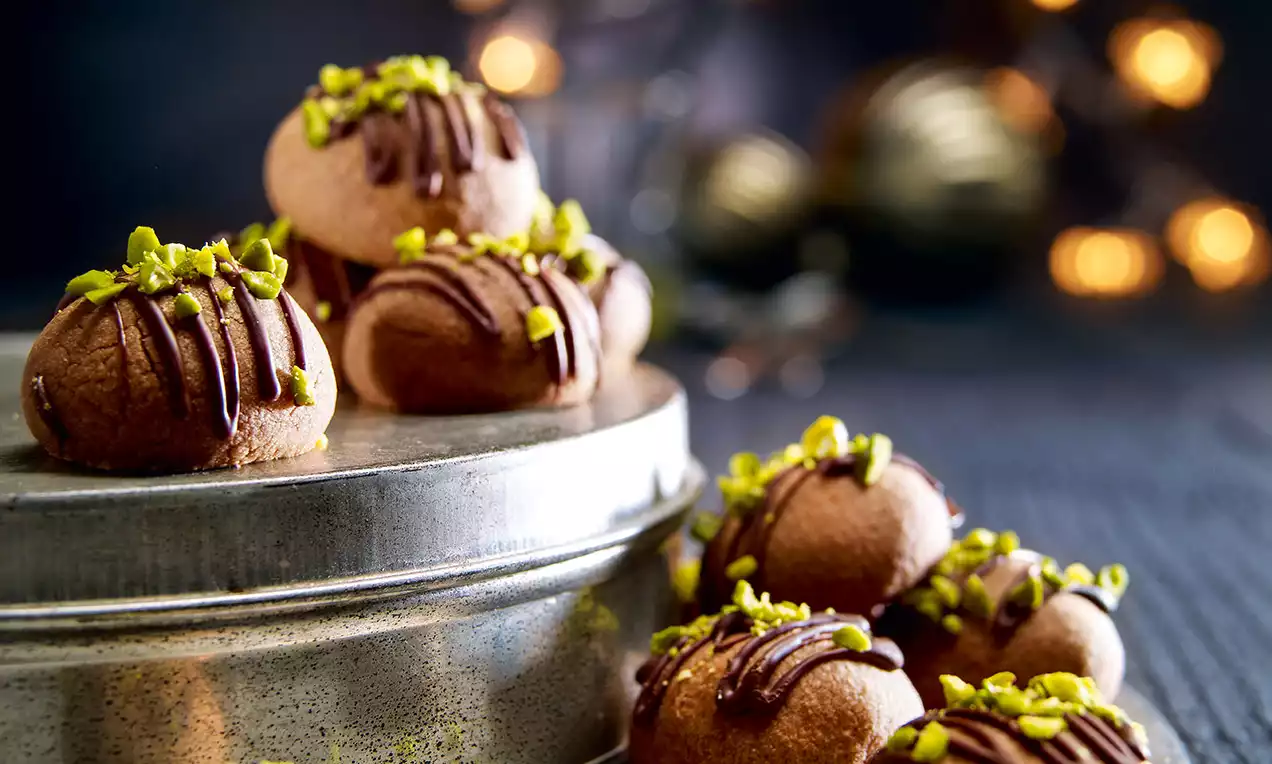Boules de nougat couleur cacao décorées de fins filets de chocolat et éclats de pistaches vertes sur une boîte métallique et un fond sombre