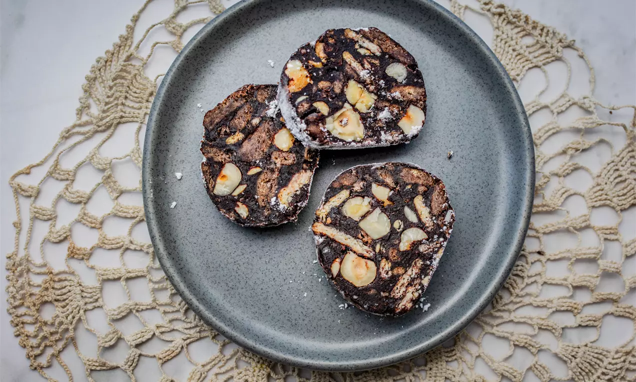 Три кръгли парчета шоколадов салам с наситено кафяв цвят, контрастни ядки и бисквити, подредени върху матово сива чиния на кремава дантела