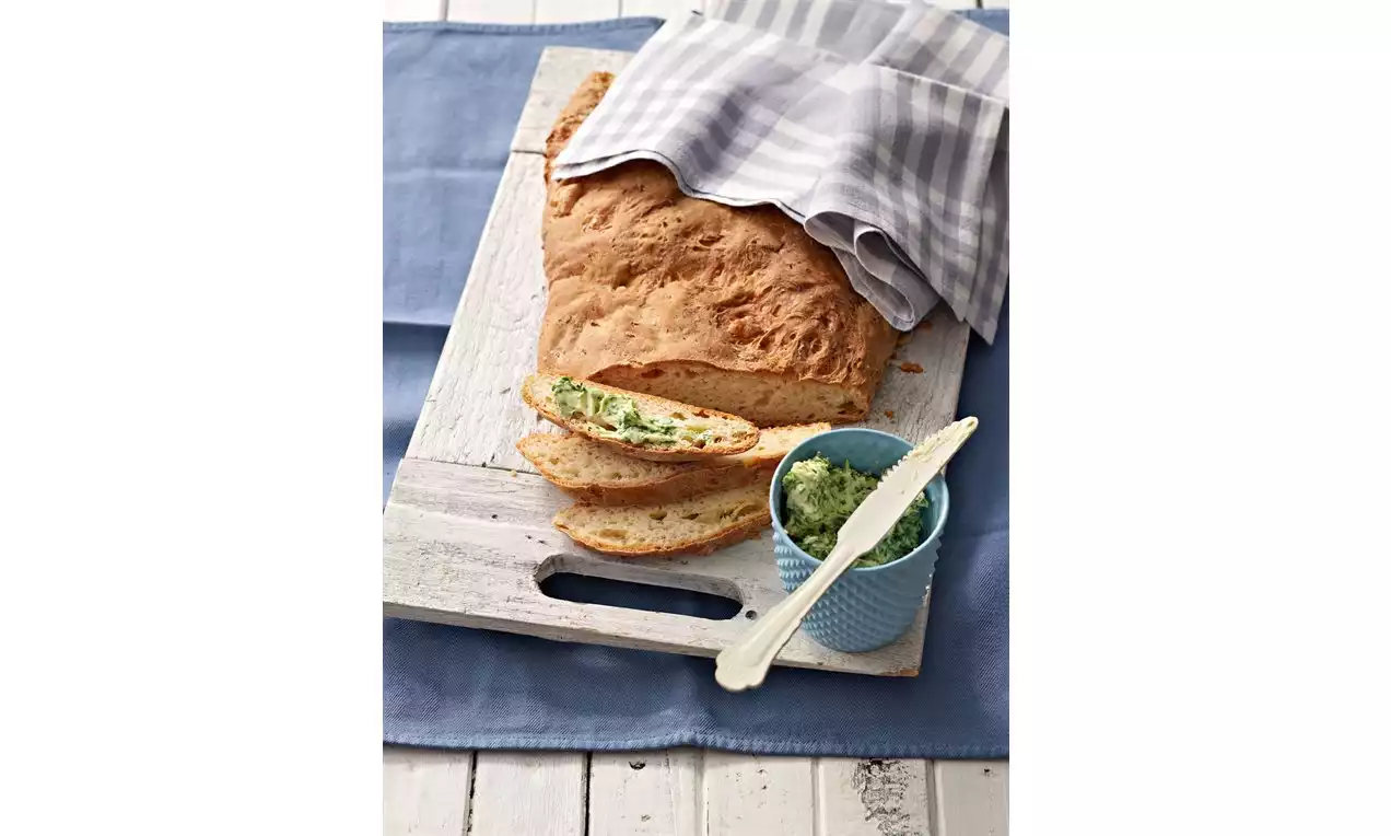 Vaaleanruskeaa, rapeapintaista leipää siivutettuna valkoisella puulevyllä, vieressä vihreää yrttivoita sinivalkoisessa keraamisessa kulhossa