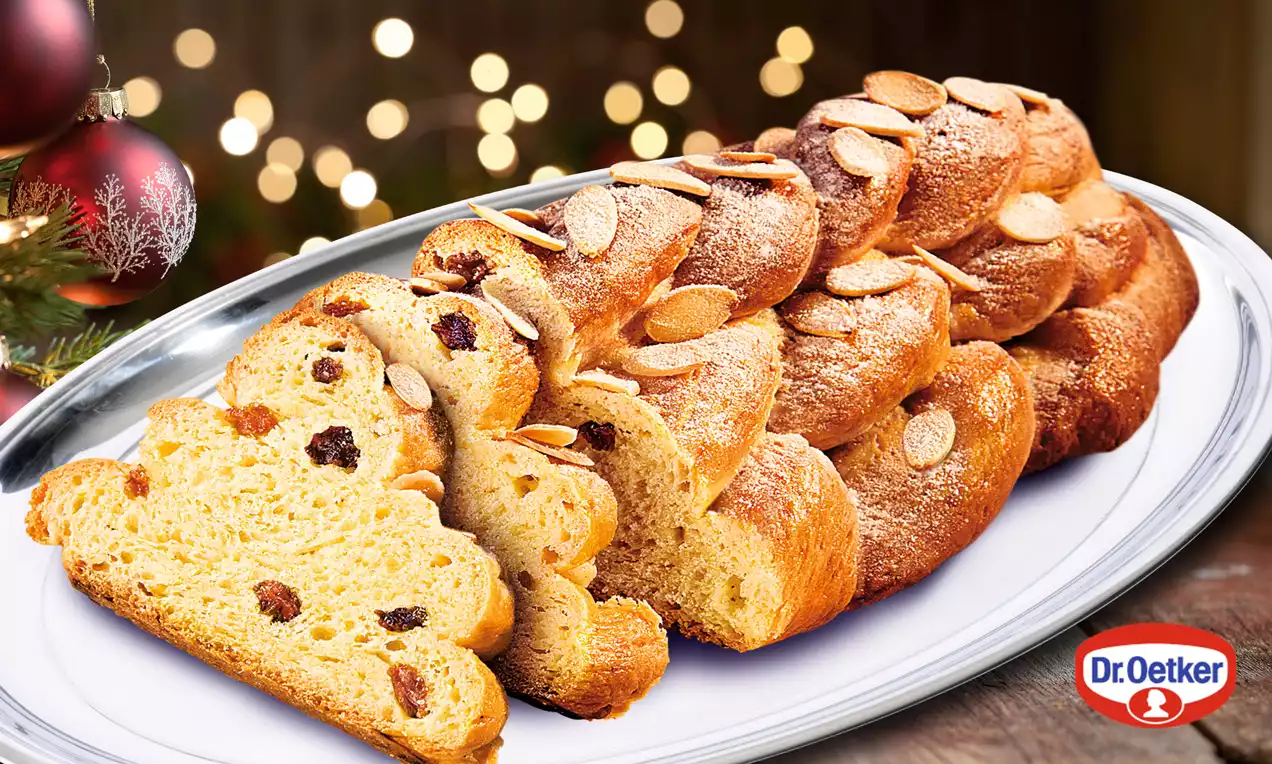 Zlatavě hnědá pletená vánočka s povrchovou posypkou mandlových plátků, uvnitř s rozinkami, na bílém oválném talíři, vánoční světýlka v pozadí