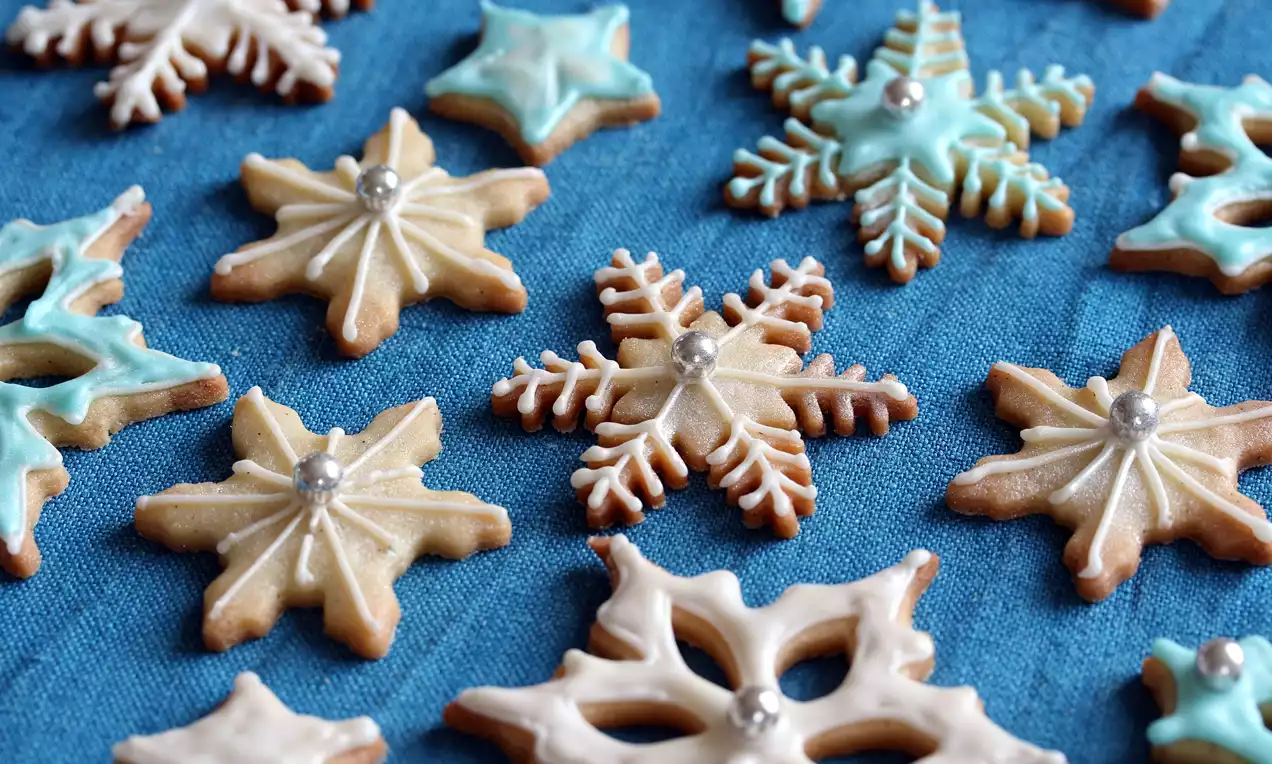 Smørkjeks formet som lyse snøkrystaller med glasur i hvitt og blått, pyntet med små sølvperler på blå tekstilbakgrunn