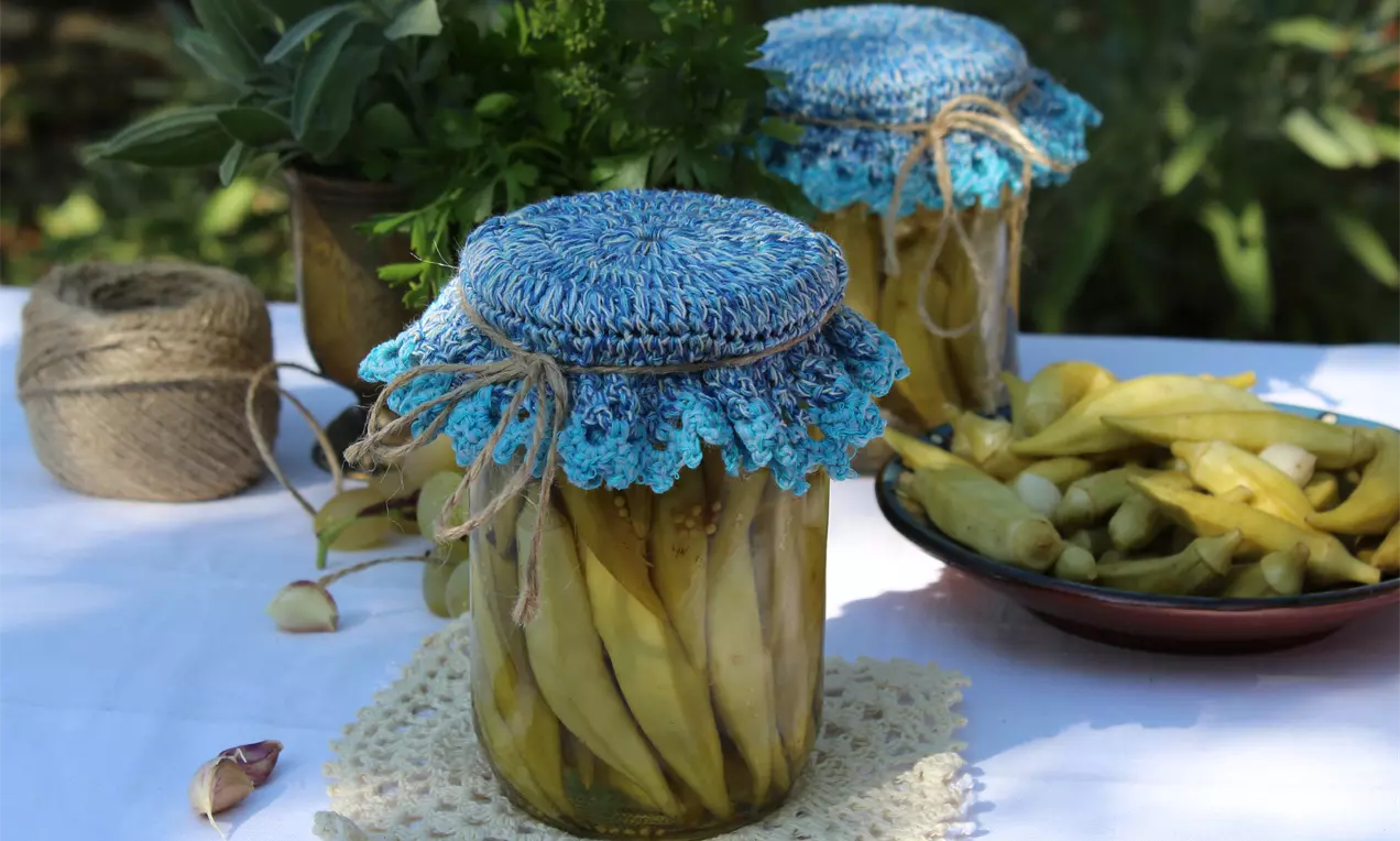 Бамята с бледозелен оттенък и нежна гладка текстура, подредена плътно в прозрачни буркани с плетени сини капачки върху бяла покривка