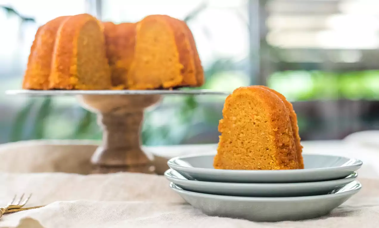 Bizcocho Bundt dorado y esponjoso con bordes curvados sobre un plato alto de pedestal y una porción servida en platos blancos apilados