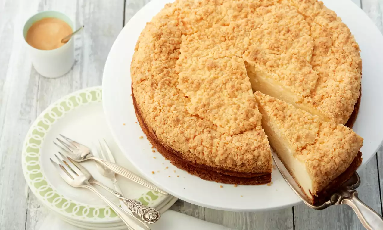 Simpler Quarkkuchen mit hellbraunen knusprigen Streuseln, rund auf weißer Tortenplatte, angeschnitten neben Gabeln und einer Tasse Kaffee