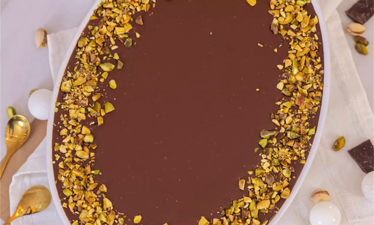 Čokoladni tiramisu u ovalnom bijelom tanjiru, tamnosmeđa površina glatka, rub ukrašen komadićima zlatnozelenih pistacija